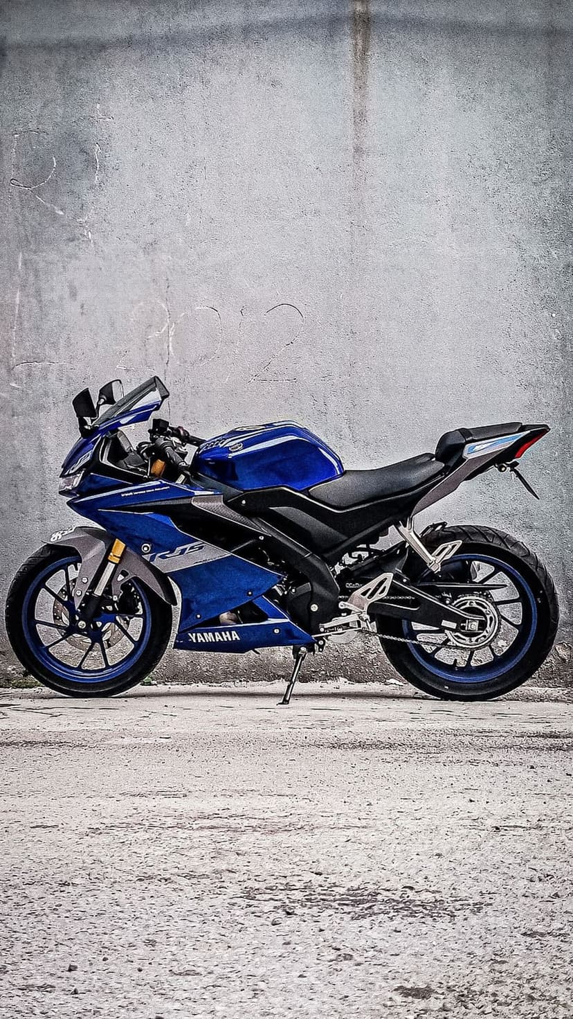 Blue Yamaha R15 motorcycle in profile view against a grey weathered concrete wall with black alloy wheels.