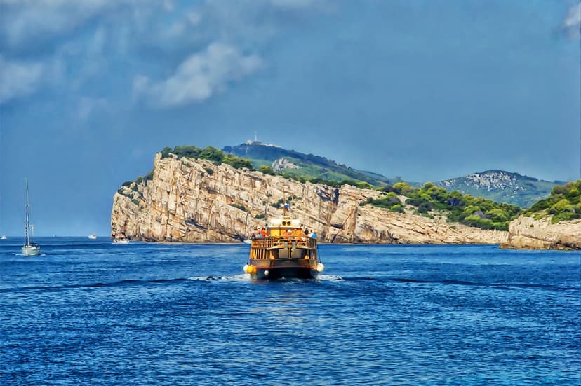 Golden tourist boat cruising past massive sunlit rocky cliffs with a white lighthouse on the distant summit.
