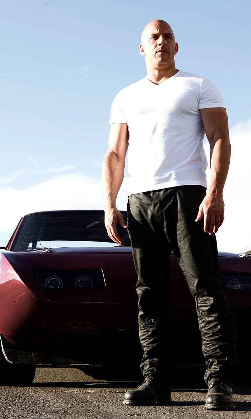 Vin Diesel in a white t-shirt leans against a dark red classic car under a clear blue sky in this portrait.