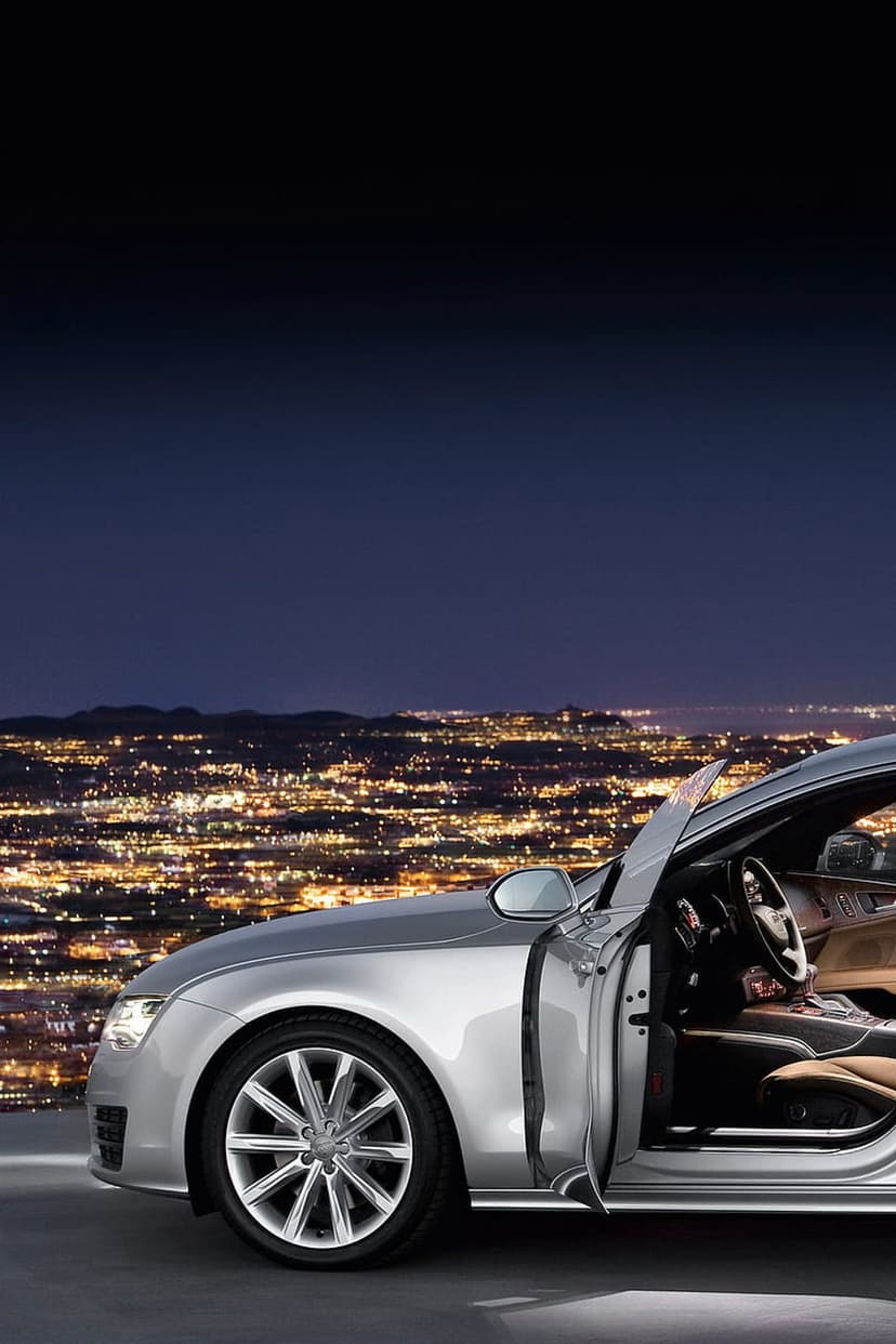 Silver Audi A7 with an open door showing tan leather interior against a backdrop of distant city lights at night.