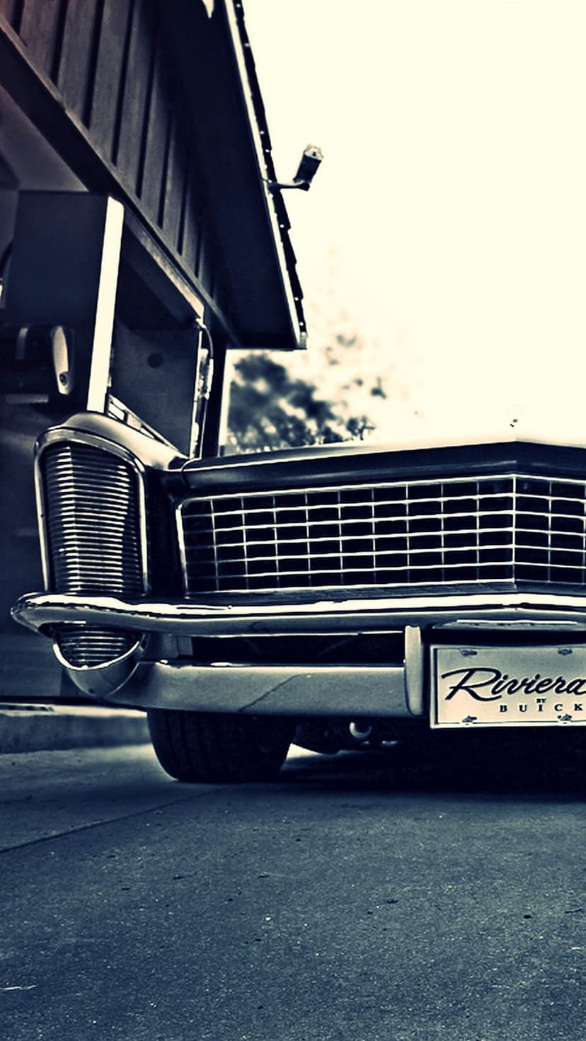 Buick Riviera chrome grill and quad headlights parked in front of a blurred urban building exterior.