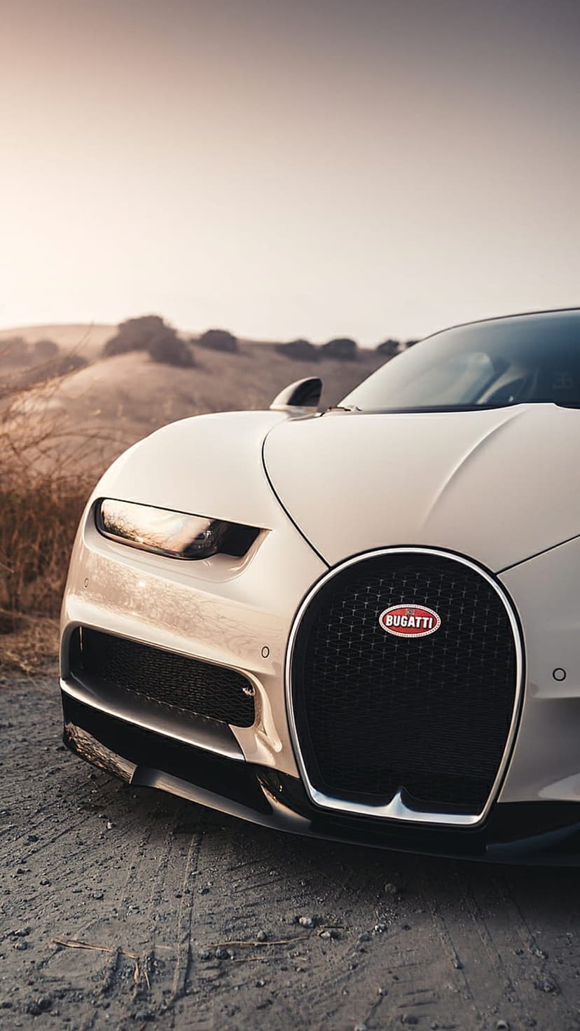 Cream Bugatti hypercar sits on a dirt road at sunset with glowing headlights and a signature horseshoe grille.