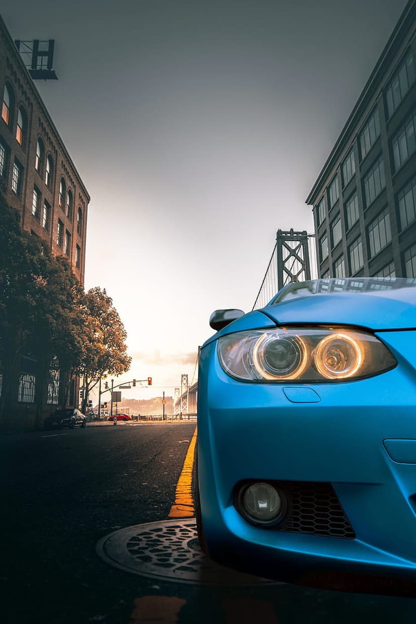 Blue sports car with glowing headlights parked on a wet city street with a suspension bridge in the distance.