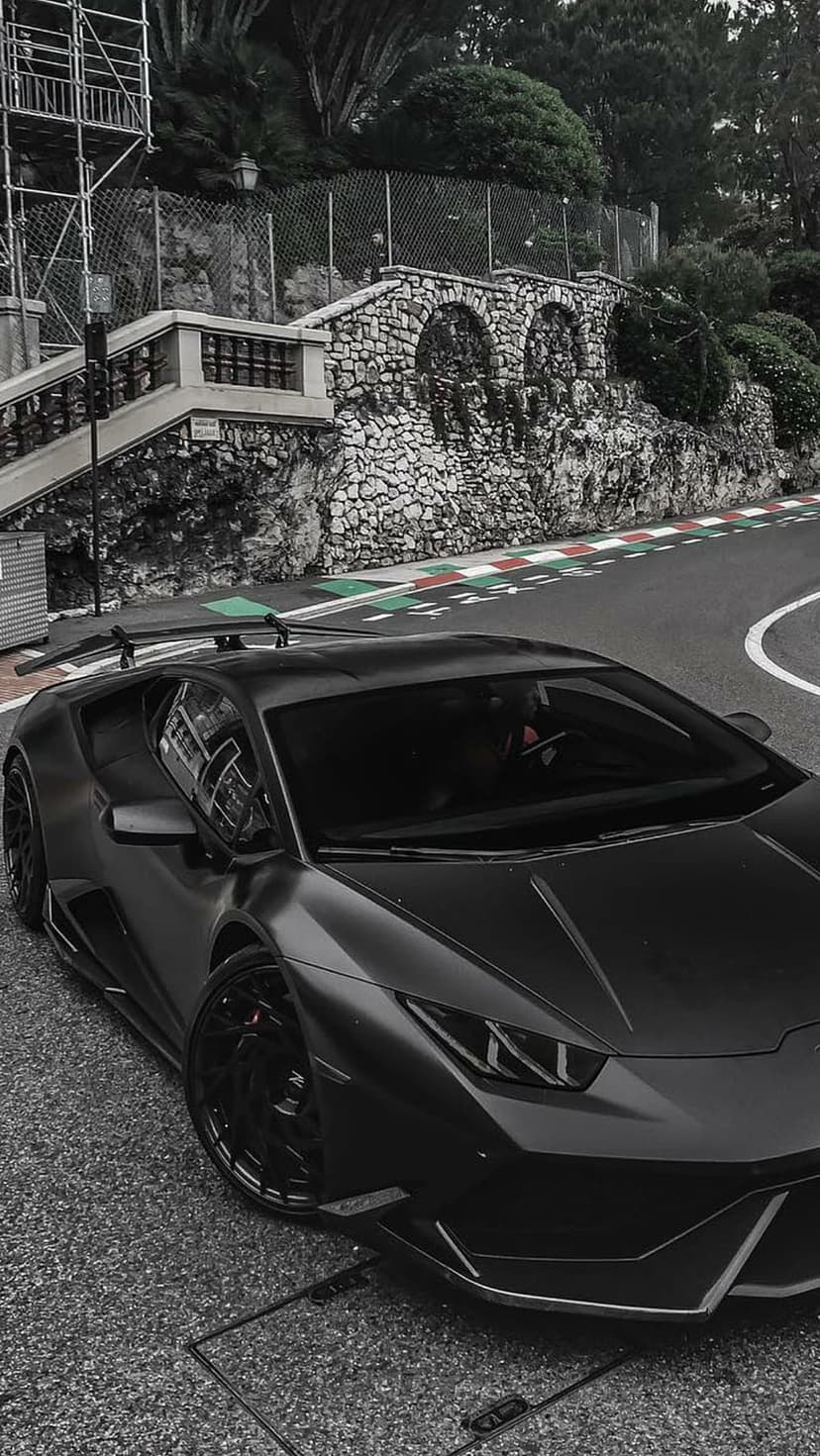 Matte black Lamborghini parked on a cobblestone Monaco street circuit with stone walls and green foliage.
