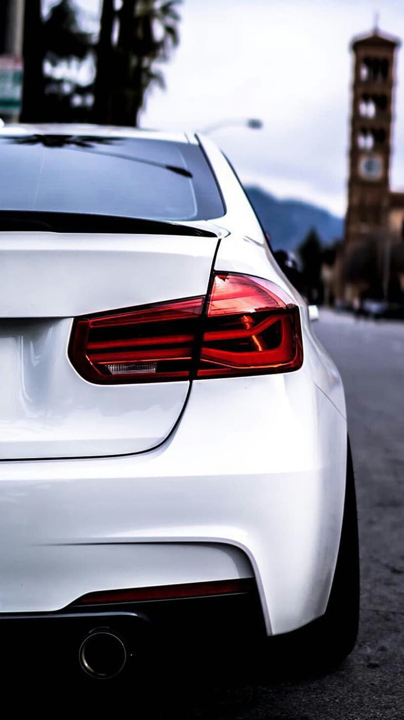 BMW L-shaped LED taillight and chrome exhaust pipe on a white car rear with a blurred urban park background.