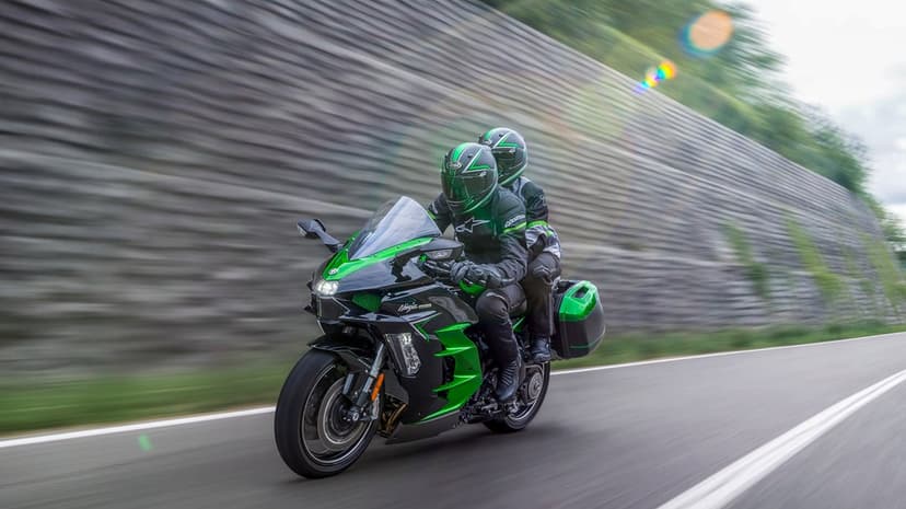 Kawasaki Ninja H2 SX motorcycle with two riders speeding past a stone wall with lens flare and motion blur.