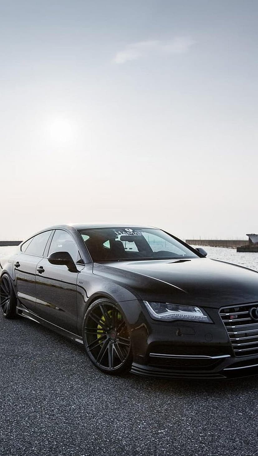 Black Audi RS7 with yellow brake calipers parked on a coastal road under a hazy sky for mobile screens.