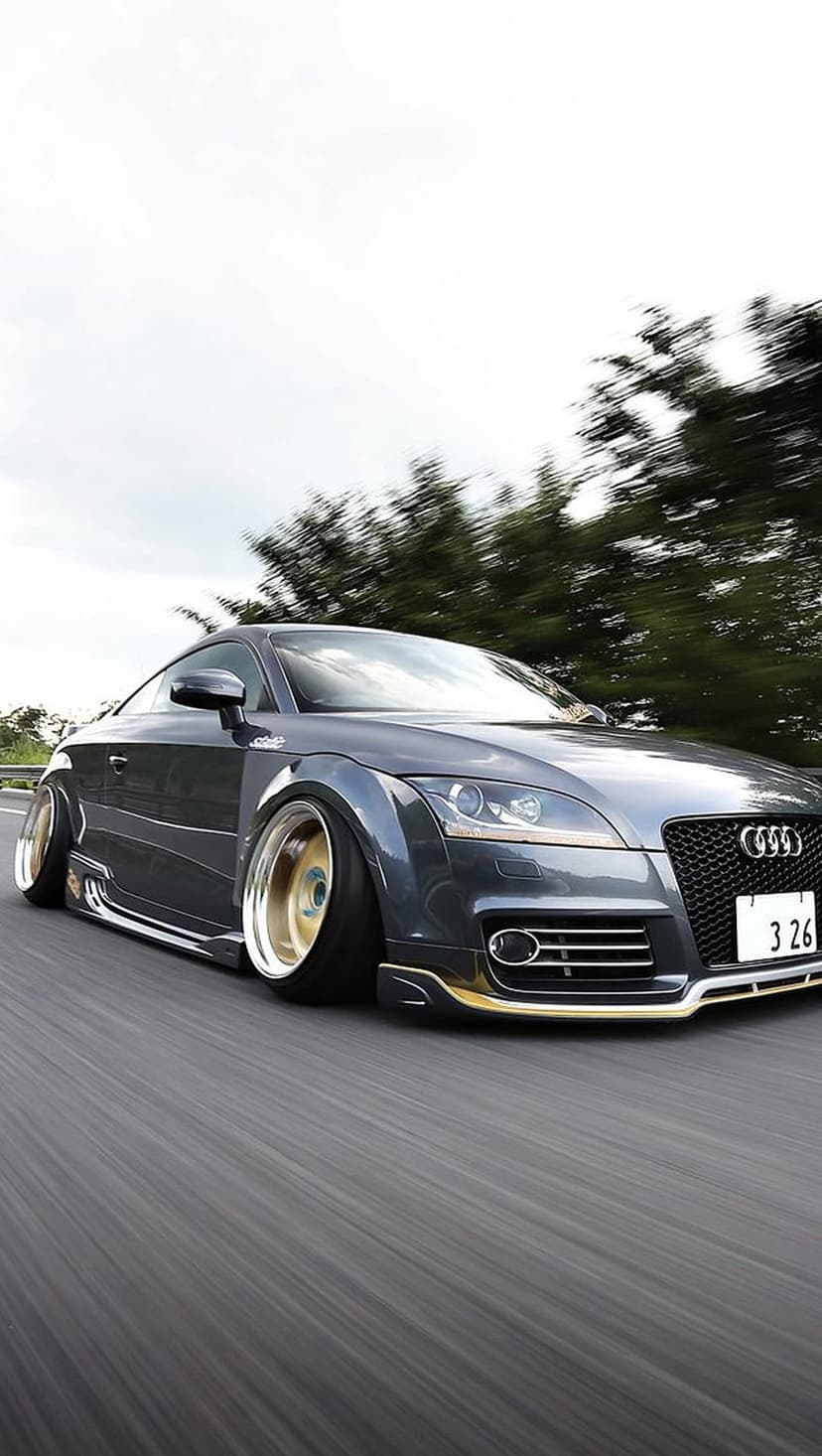 Dark gray Audi TT with gold rims driving fast on a paved road with blurred green trees in the background.