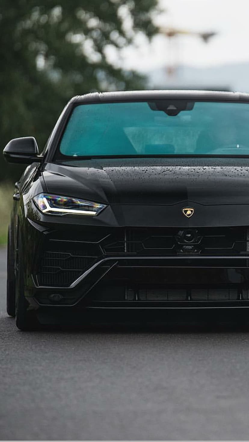 Black Lamborghini Urus with LED headlights and raindrops on its hood parked on a dark, wet asphalt road.