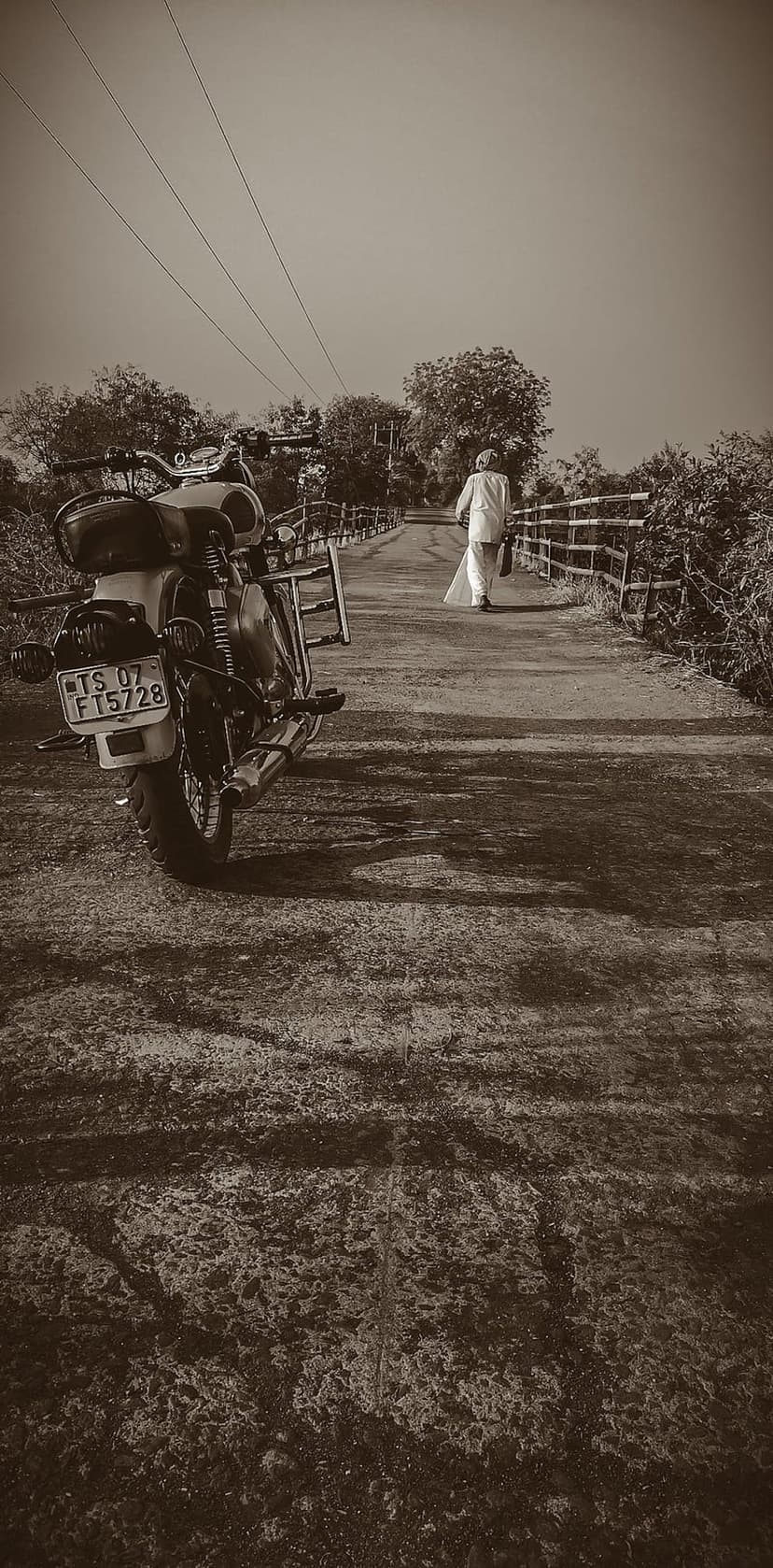 Motorcycle parked on a rustic bridge while a person in white walks toward a misty forest in sepia tones.