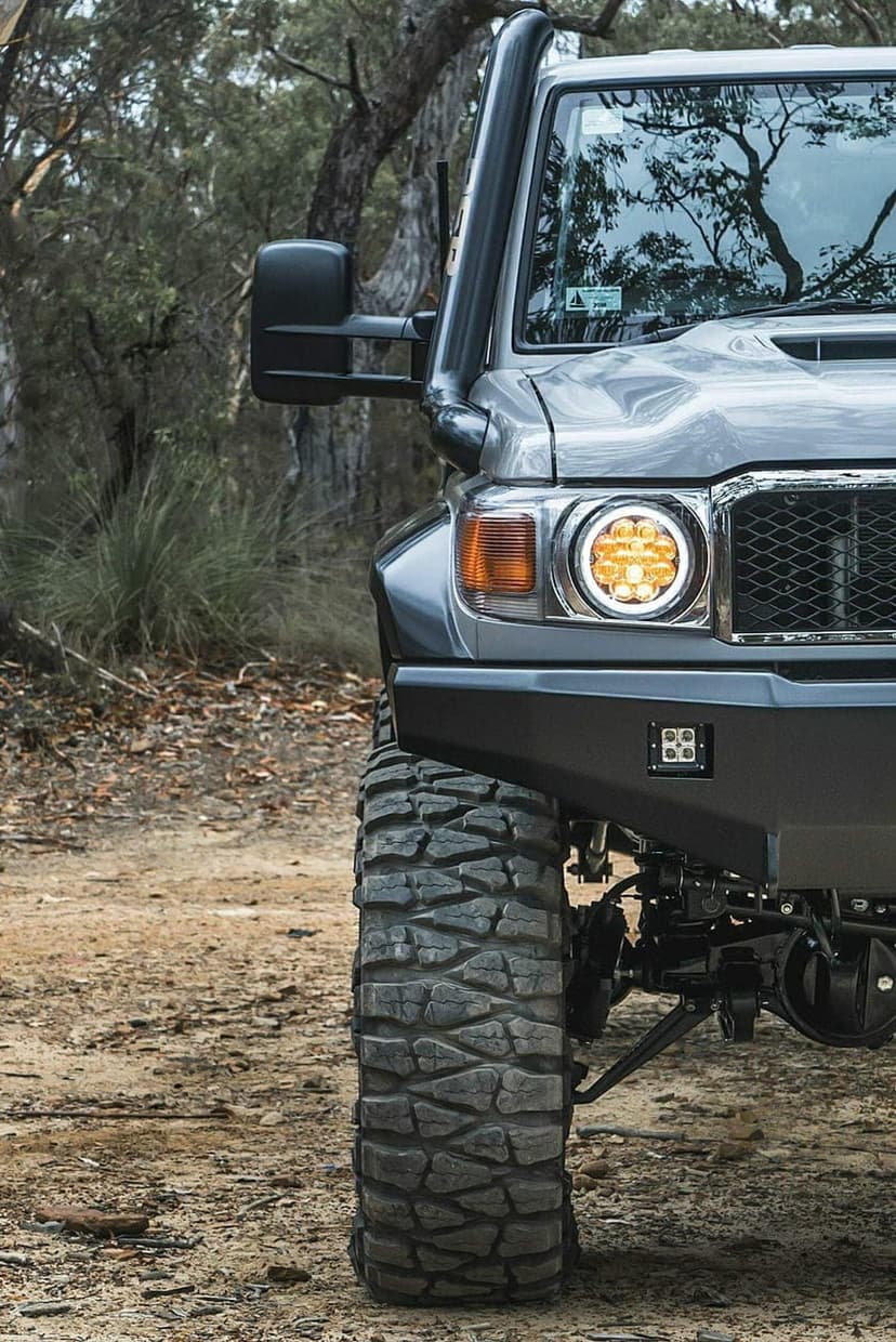 Gray 4x4 truck with a snorkel and aggressive mud tires parked on a dirt forest path between tall trees.