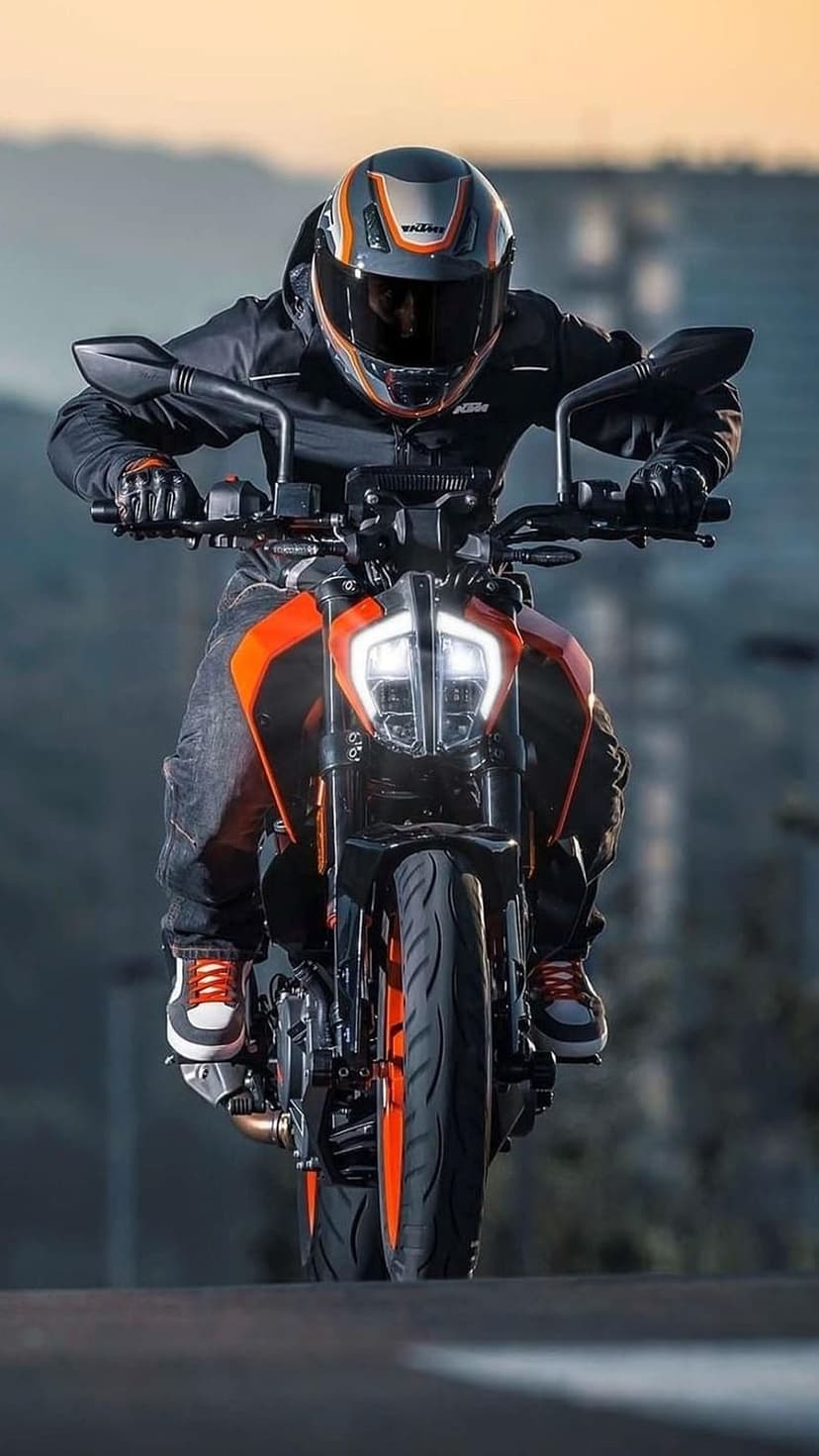 Motorcyclist in black gear and orange sneakers rides a KTM bike with glowing headlights on a blurred city road.
