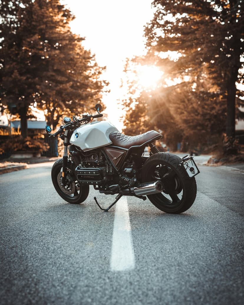Cafe racer motorcycle with a white tank and brown seat parked on an asphalt road during a golden sunset with lens flare.