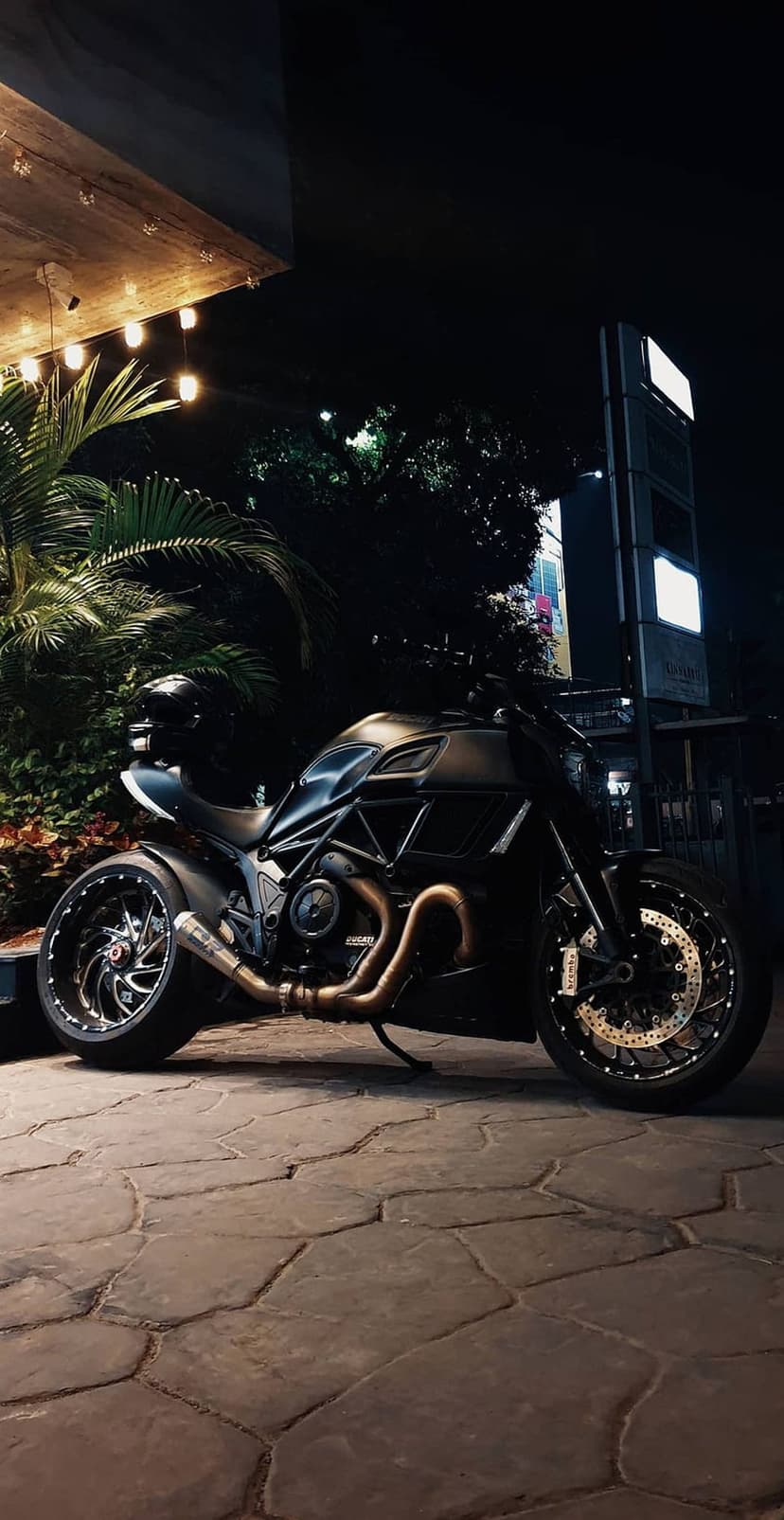 Black Ducati motorcycle on a stone patio at night under warm string lights with palm leaves in the background.