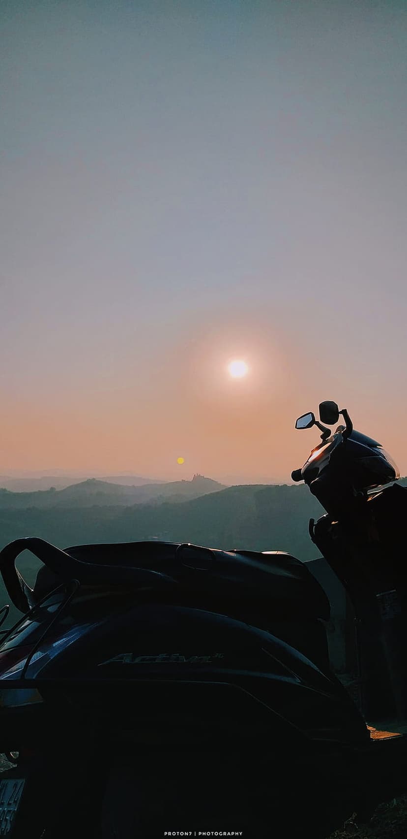 Vintage scooter parked on a grassy ridge overlooking a misty mountain valley during a golden orange sunset.