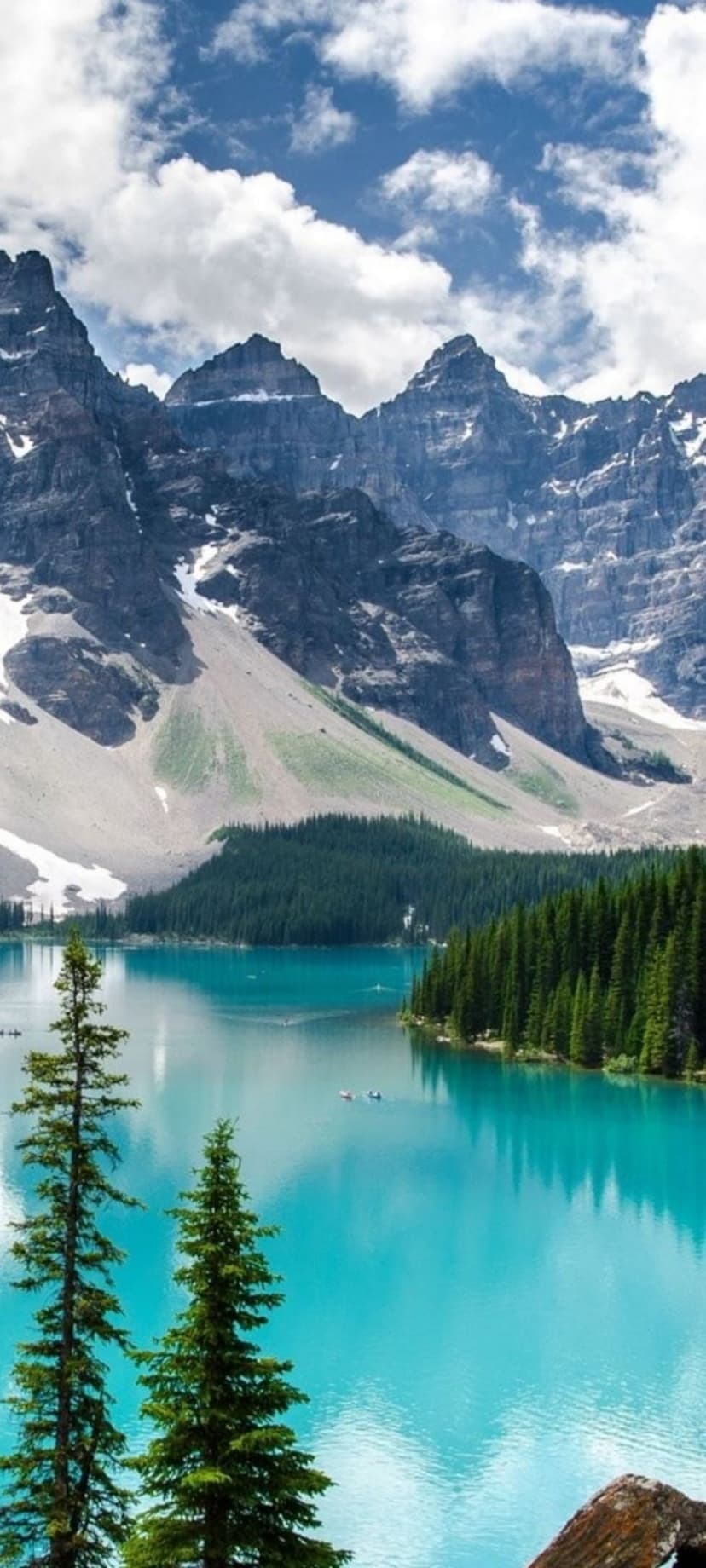 Turquoise glacial lake surrounded by evergreen forests and jagged snow-covered mountains under a bright blue sky.