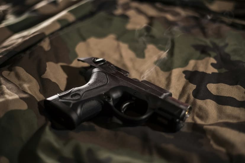 Black semi-automatic pistol with rising smoke resting on a green and brown camouflage fabric background.