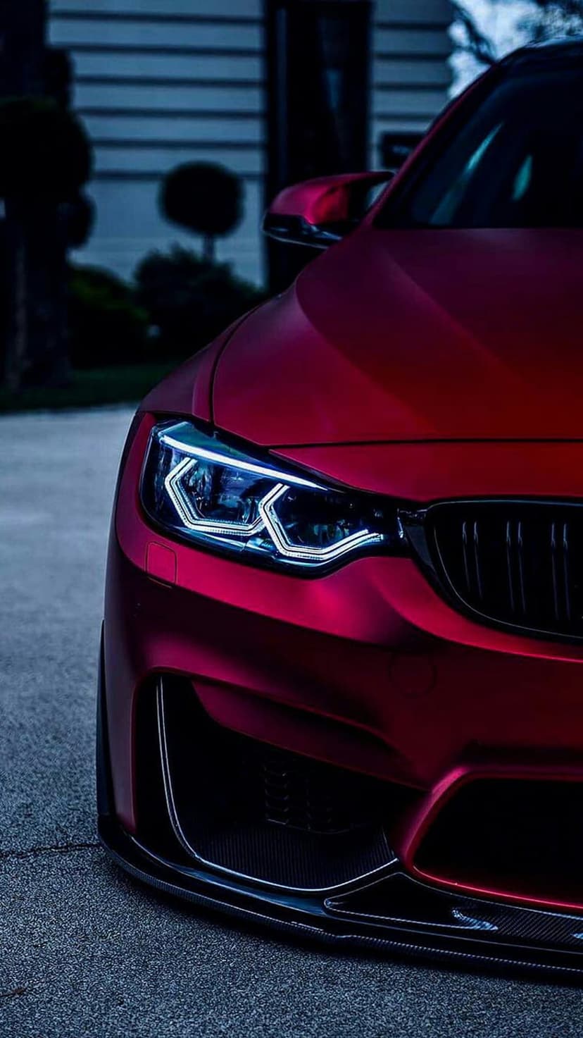 Crimson red sports car headlights glow with bright white LED beams against a dark matte front bumper at dusk.