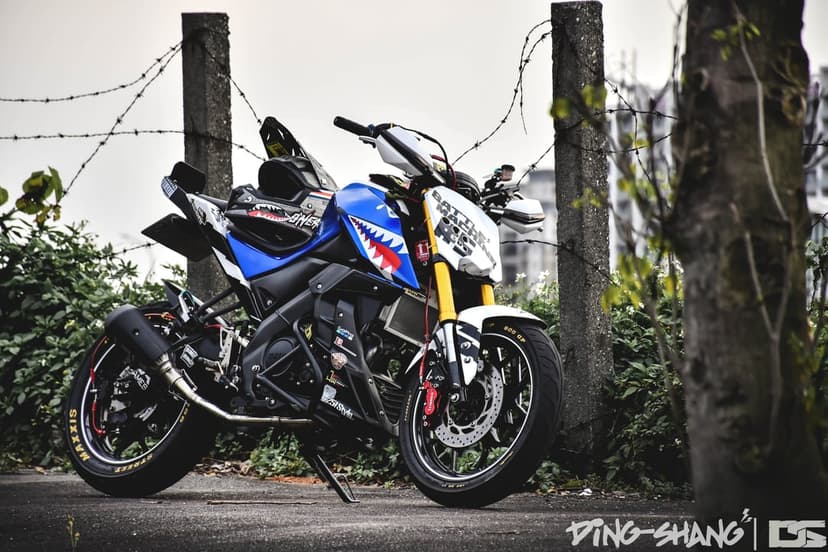 Yamaha motorcycle with blue and white shark teeth graphics and yellow suspension near a barbed wire fence.