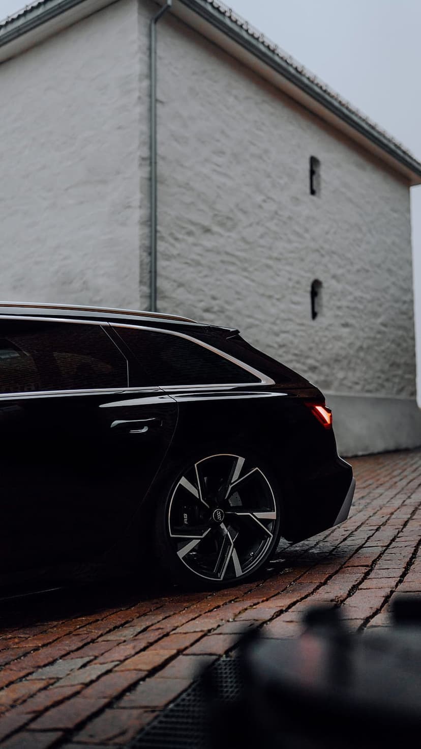Black Audi RS6 Avant station wagon parked on rain-slicked brick street next to a white textured building.