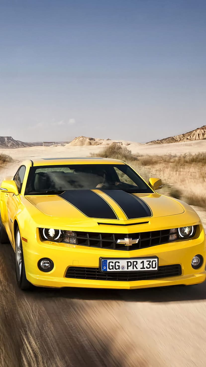 Yellow Chevrolet Camaro with black stripes kicks up dust on a desert road with mountains under a clear blue sky.