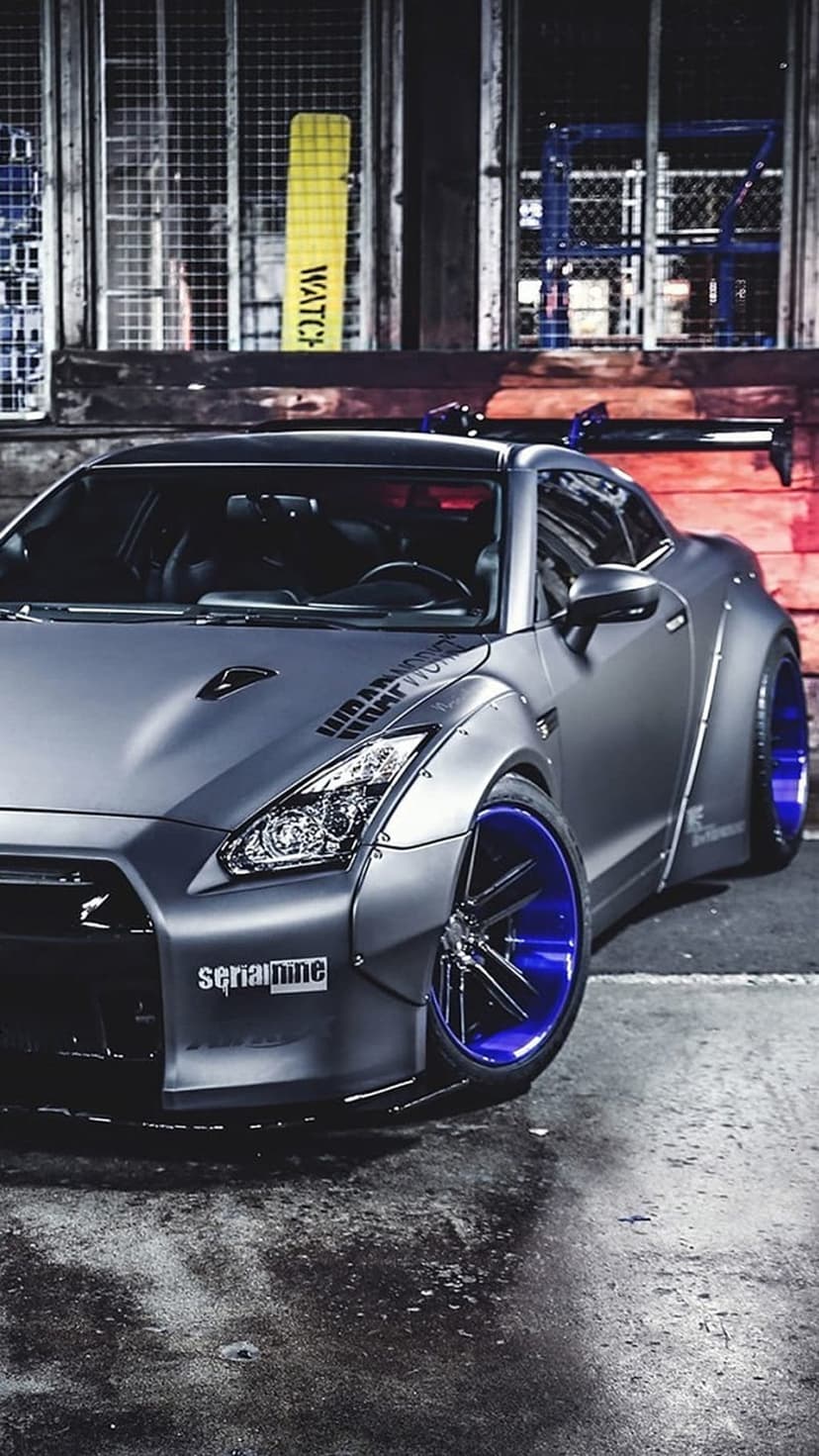 Nissan GT-R with a matte grey widebody kit and bright blue rims parked beside a rustic brick wall and metal fence.
