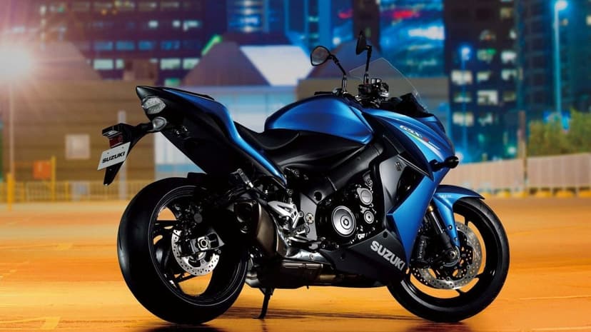 Blue Suzuki GSX-S1000F sports motorcycle parked on a warm orange ground against a blurred city night background.