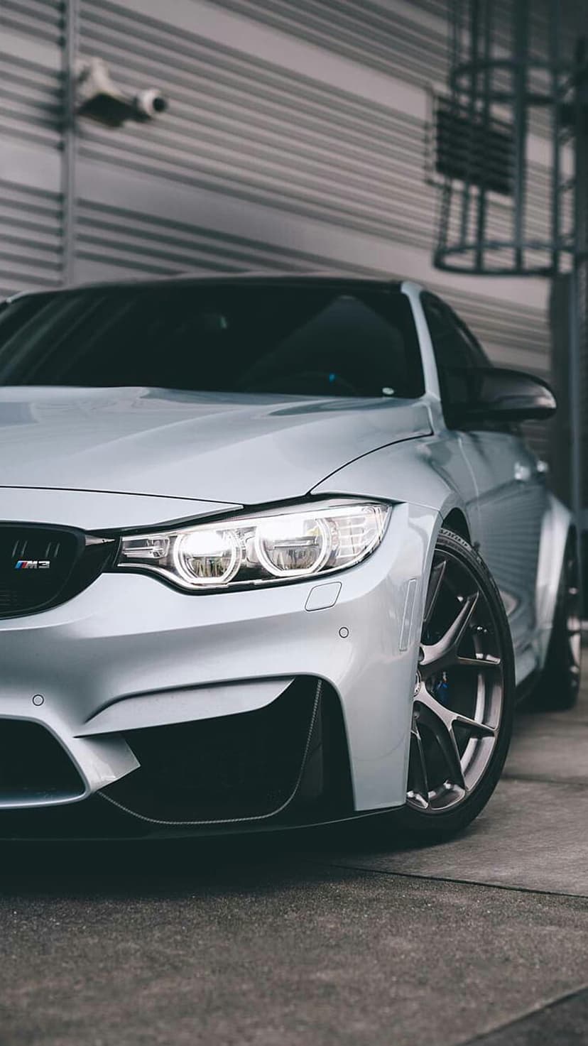 Silver BMW M3 front bumper and LED headlight shown at a low angle against a textured metallic steel wall.