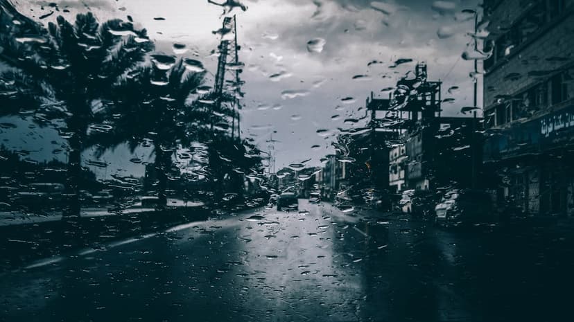 Water droplets on a glass window pane overlook a blurred city street with buildings under a grey overcast sky.