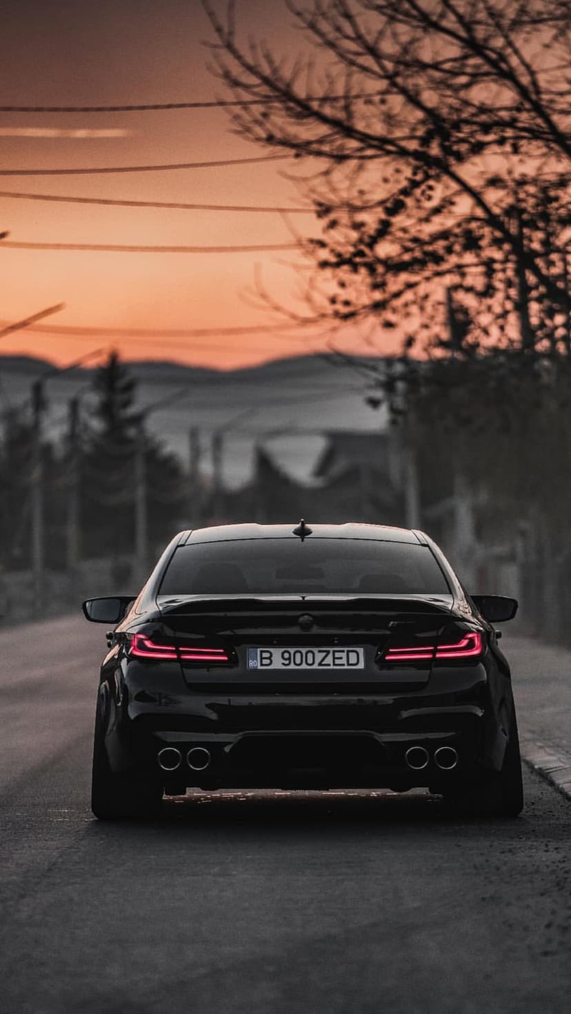 BMW M5 sedan rear view parked on asphalt under an orange sunset sky with glowing taillights and silhouetted trees.