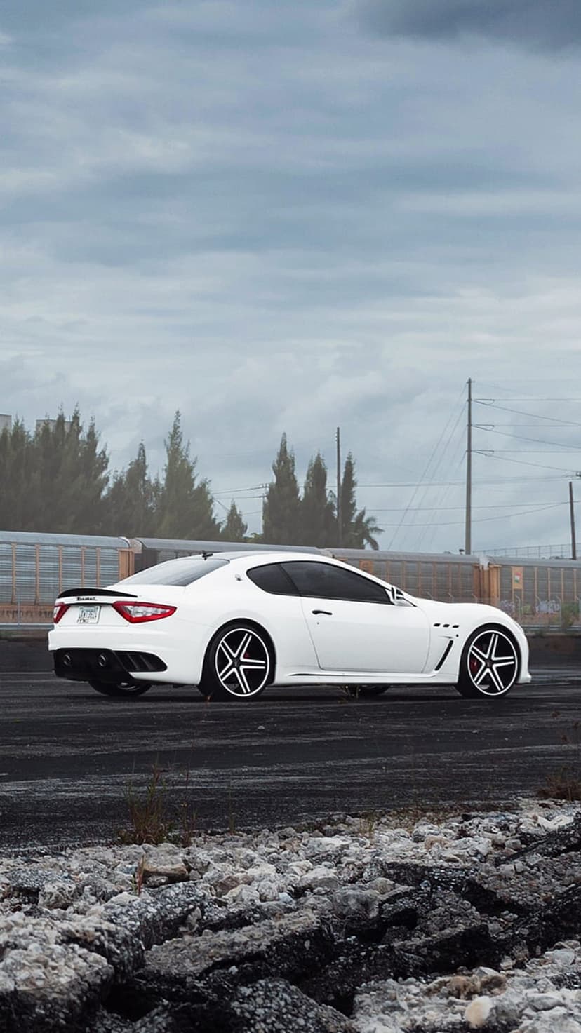 White Maserati GranTurismo side profile on asphalt with custom wheels under a dark, stormy, overcast sky.