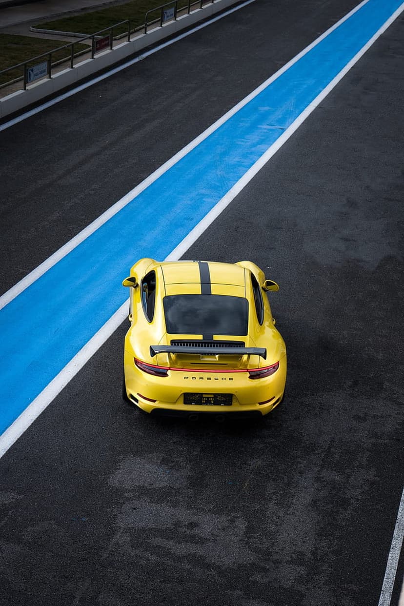 Yellow Porsche 911 with black stripes drives on a dark asphalt race track with blue and white painted lines.