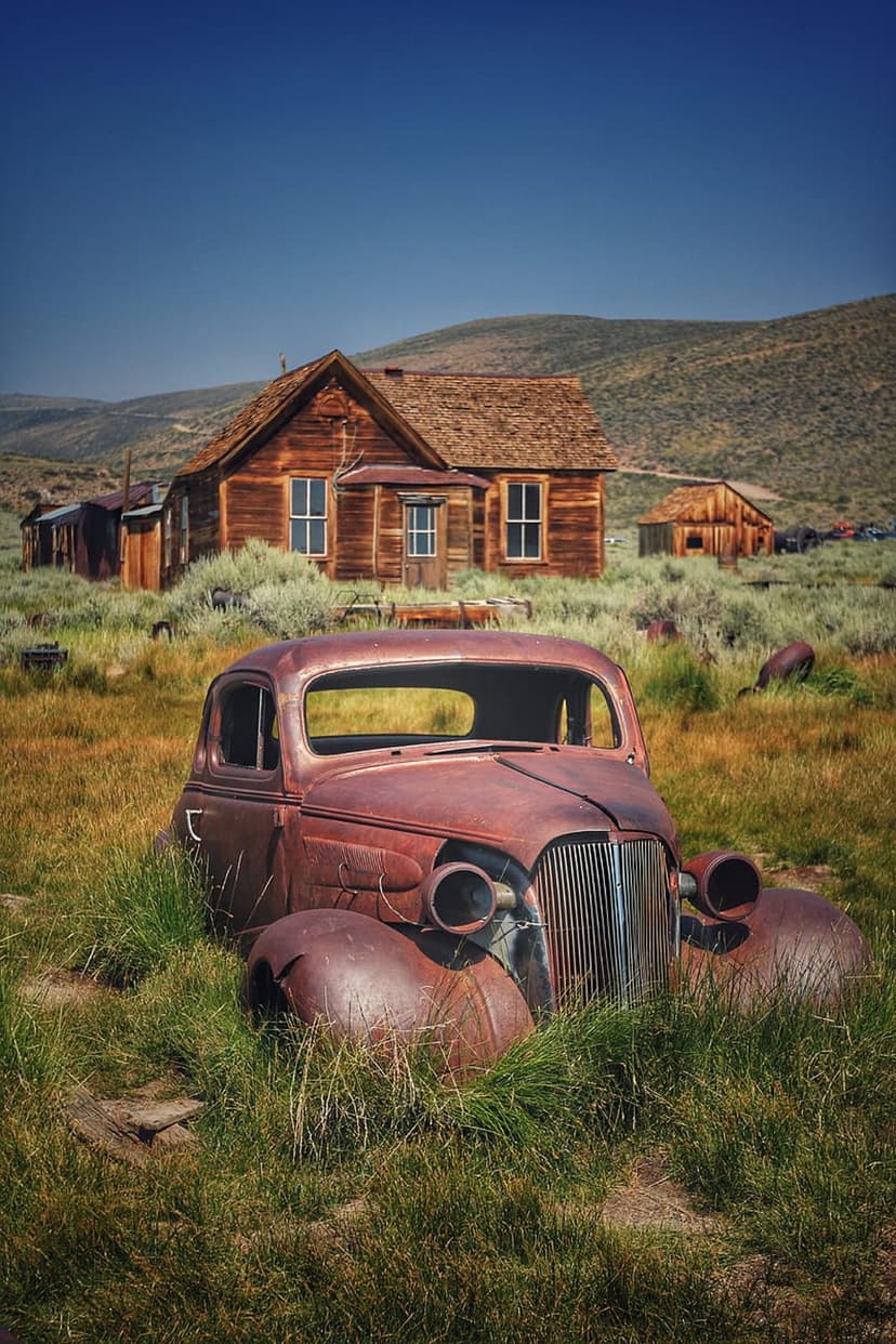 Rusty vintage car sits in tall grass before wooden ghost town buildings under a clear blue sky in rural hills.
