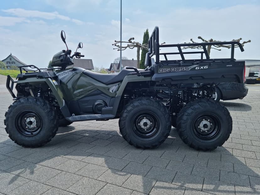 Side view of a green Polaris 6x6 ATV with a black cargo bed and six large off-road tires on a paved driveway.