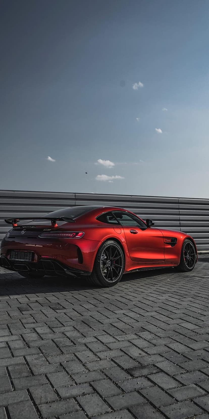 Red Mercedes-AMG GT R with a large rear wing parked on brick pavers against a corrugated metal wall and blue sky.
