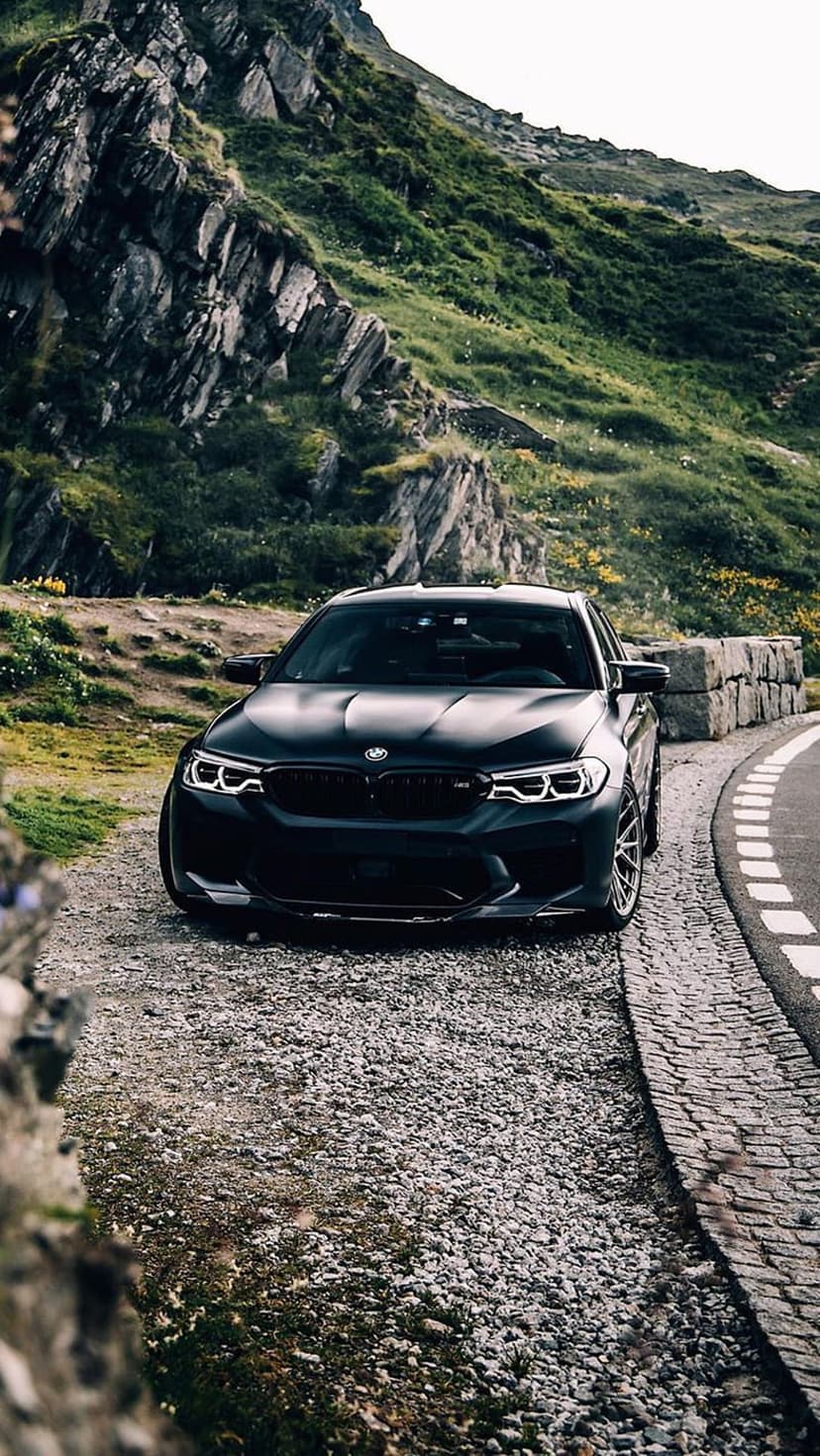 Black BMW M5 parked on a rocky mountain road with green slopes and yellow flowers in the background.
