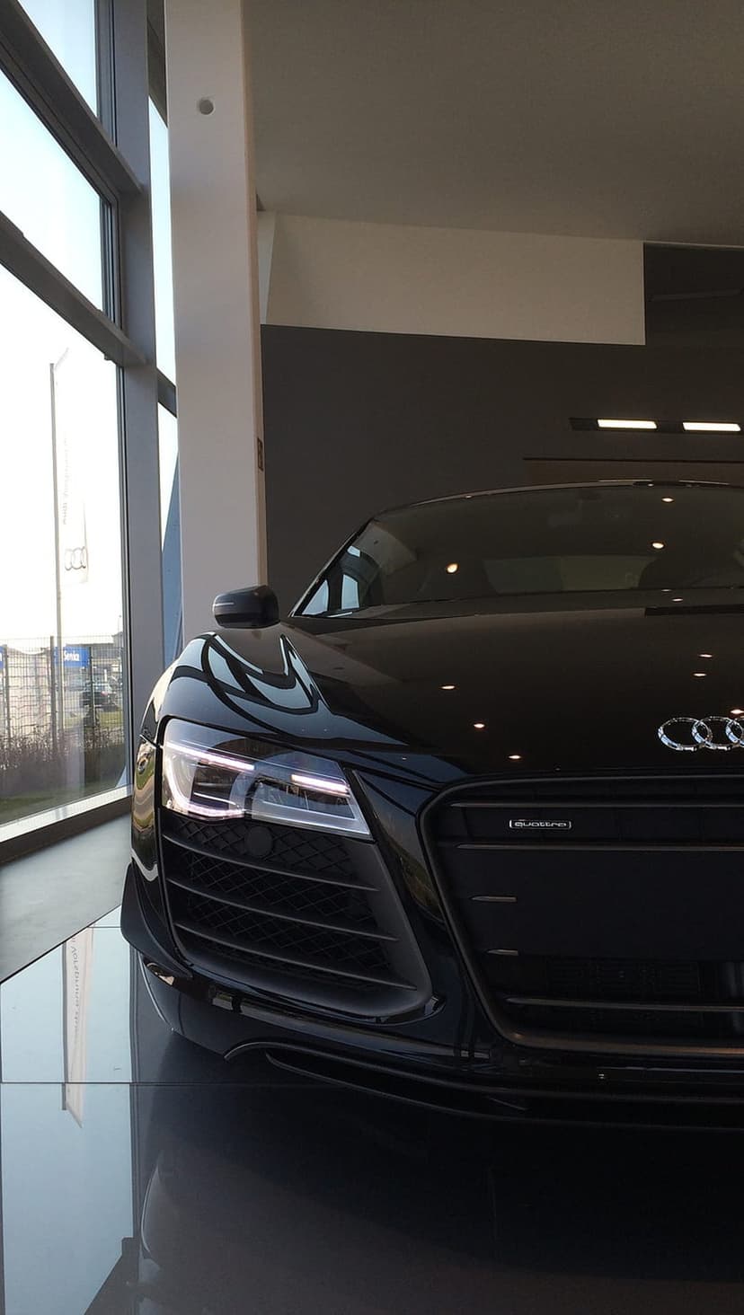 Black Audi R8 with glowing LED headlights parked on a reflective floor inside a sunlit modern car showroom.