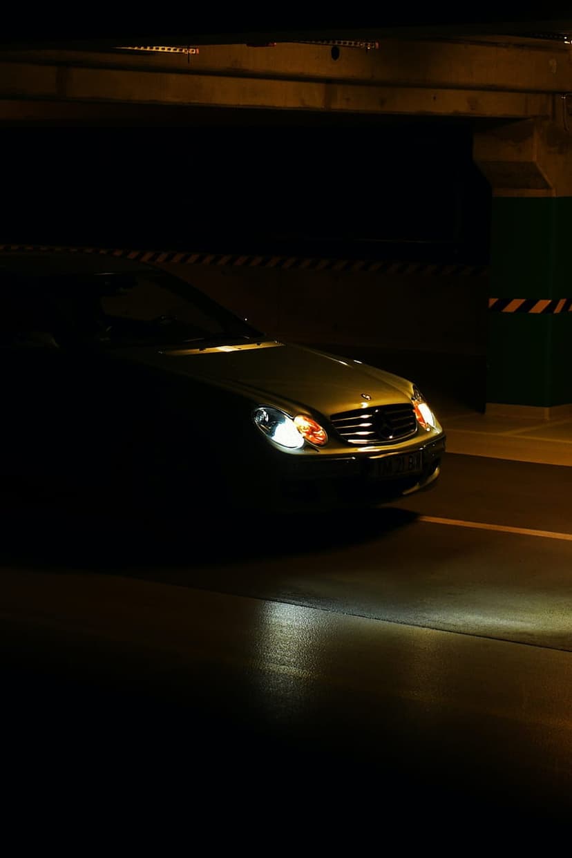 Luxury car headlights cast a bright white glow onto wet concrete pavement inside a dark underground parking garage.
