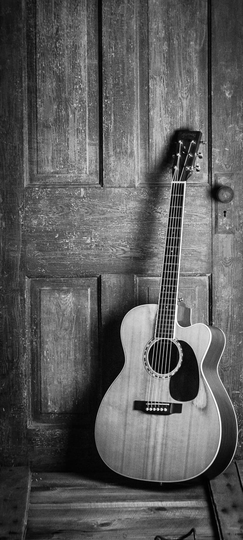 Acoustic guitar leaning against a textured wooden door with vertical planks in a black and white photograph.