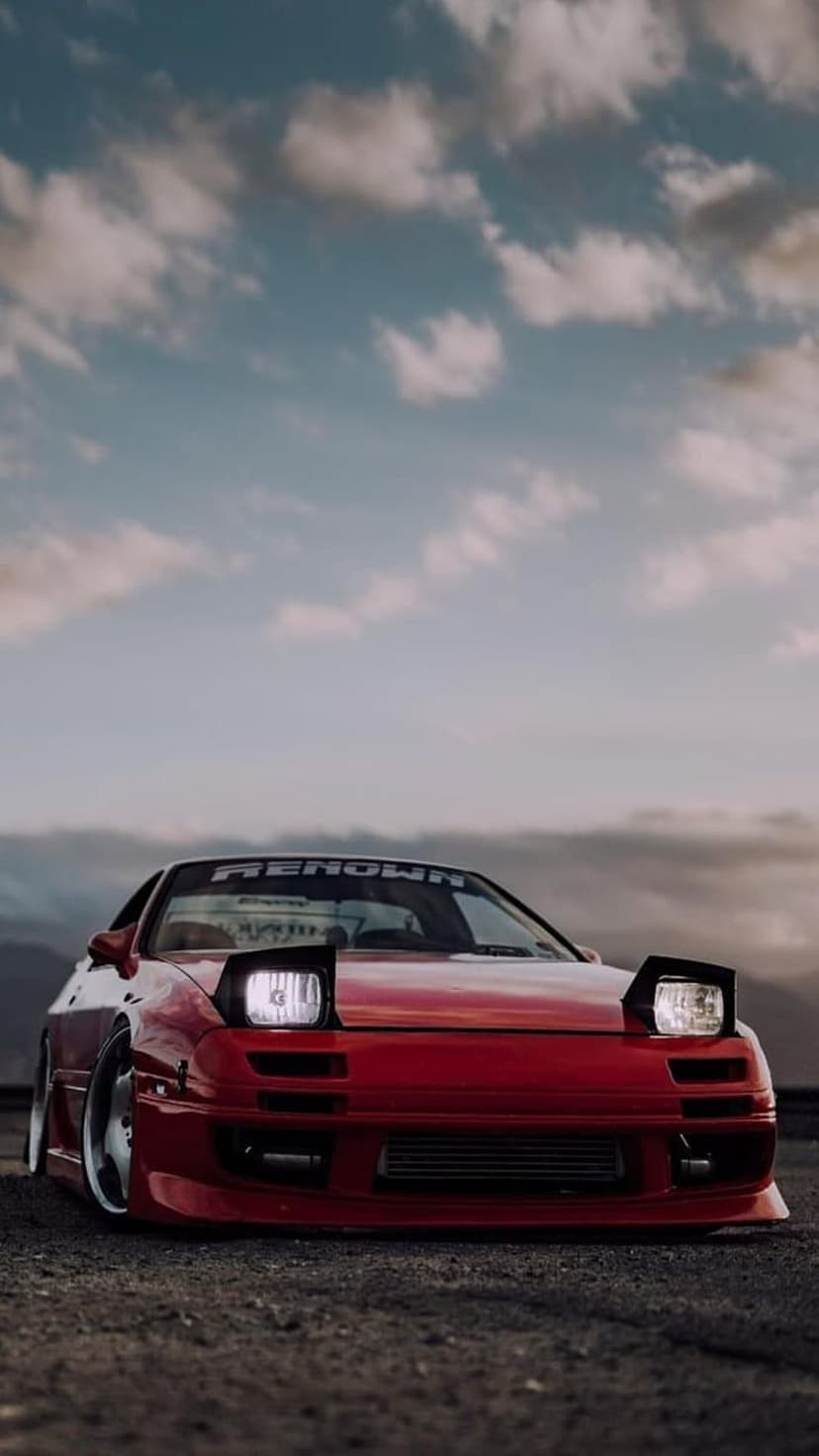Red Mazda RX-7 FC sports car with lit pop-up headlights and Renown banner parked on gravel under a cloudy sky.