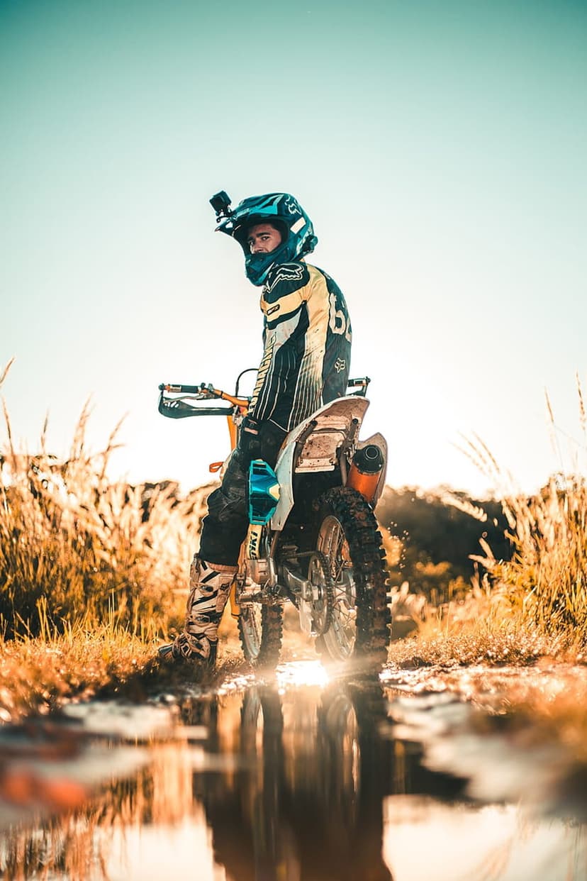 Motocross rider in full gear standing on a dirt bike over a reflective puddle in a grassy field at sunset.