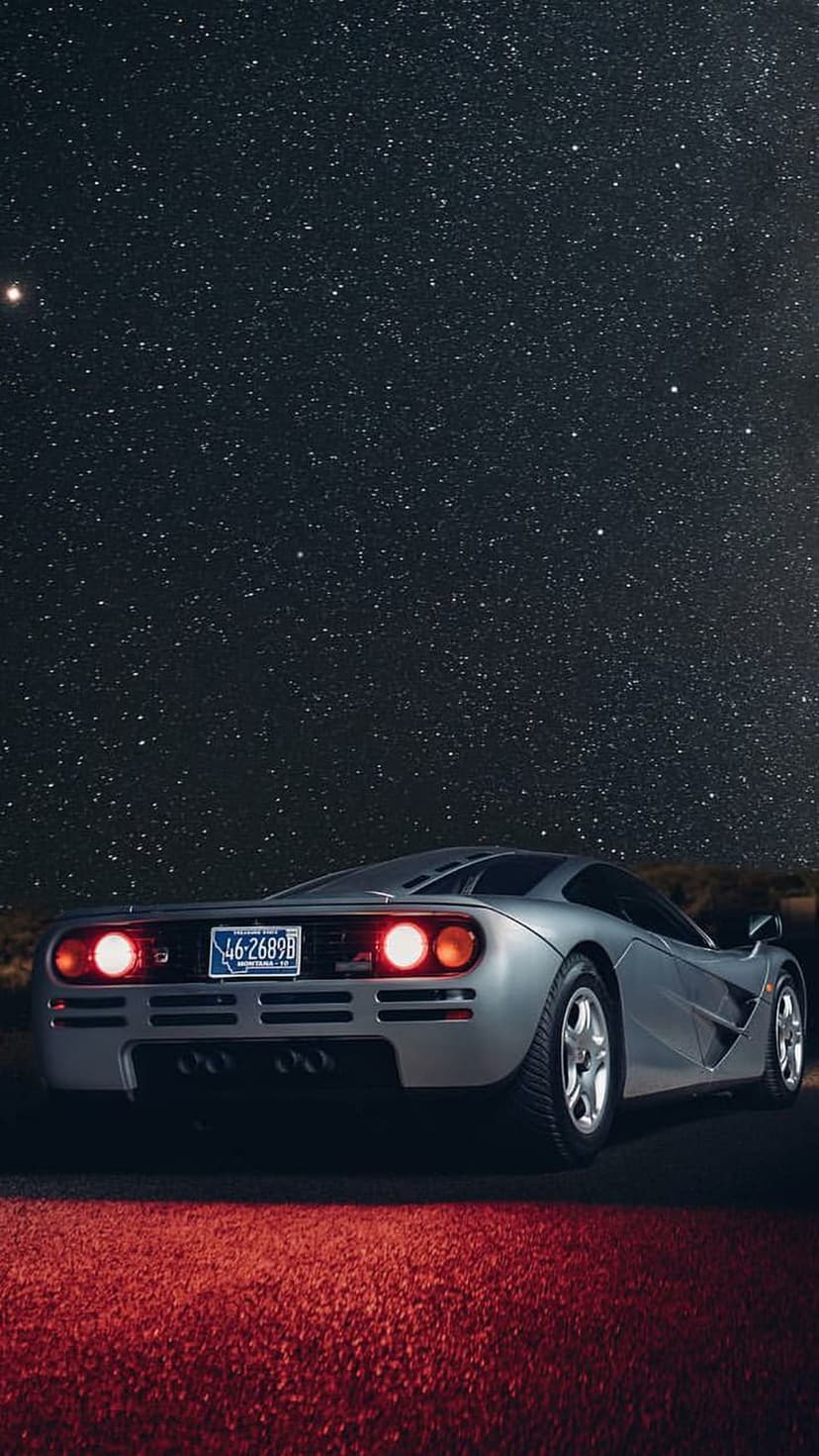McLaren F1 supercar with glowing red taillights parked on a dark road beneath a vast starry night sky.