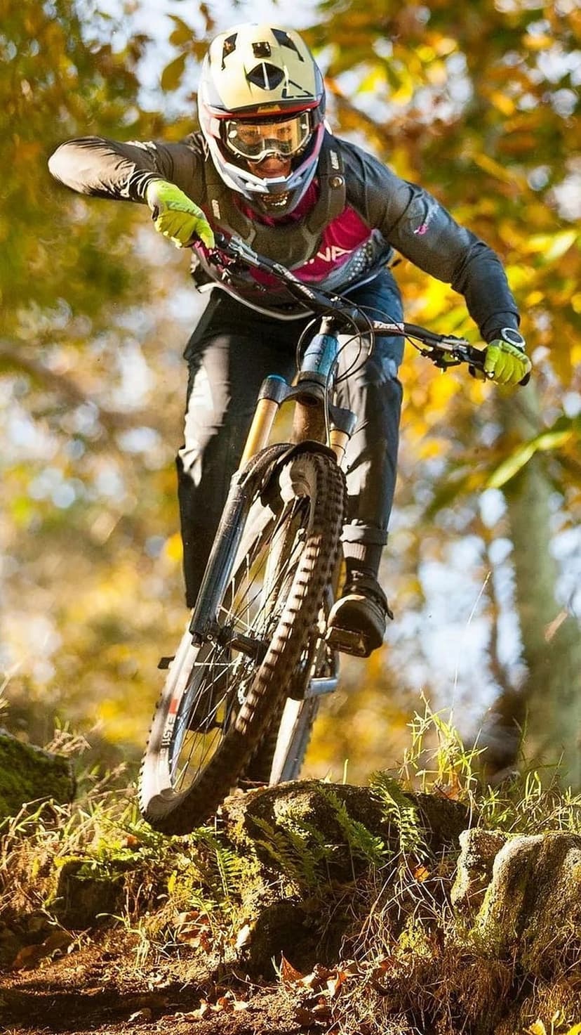 Mountain biker in full gear leans into a rocky forest turn with autumn leaves, ideal for a vertical screen.