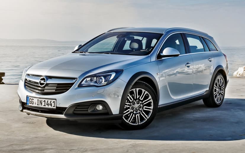 Silver Opel Insignia Country Tourer wagon parked on a concrete pier near a calm blue sea under a bright, hazy sky.