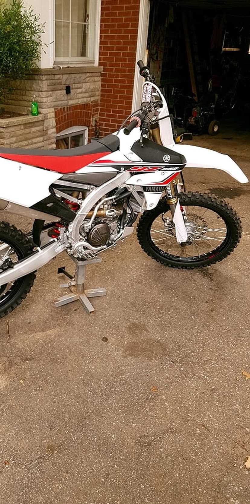 Yamaha dirt bike with red accents sits on a silver metal stand in front of a brick house and garage area.
