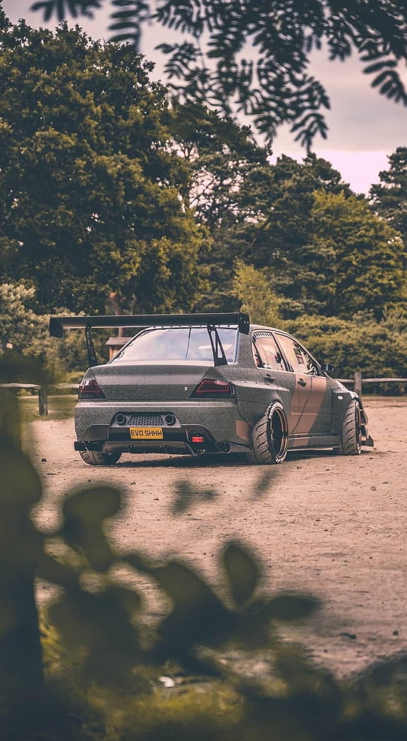 Mitsubishi Lancer Evolution with a large rear wing and widebody fenders parked on a dirt road in a green forest.