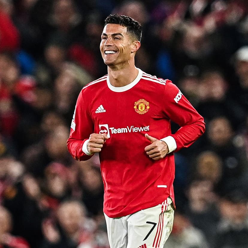 Cristiano Ronaldo smiles on a grass pitch wearing a red Manchester United long-sleeved kit and white shorts.
