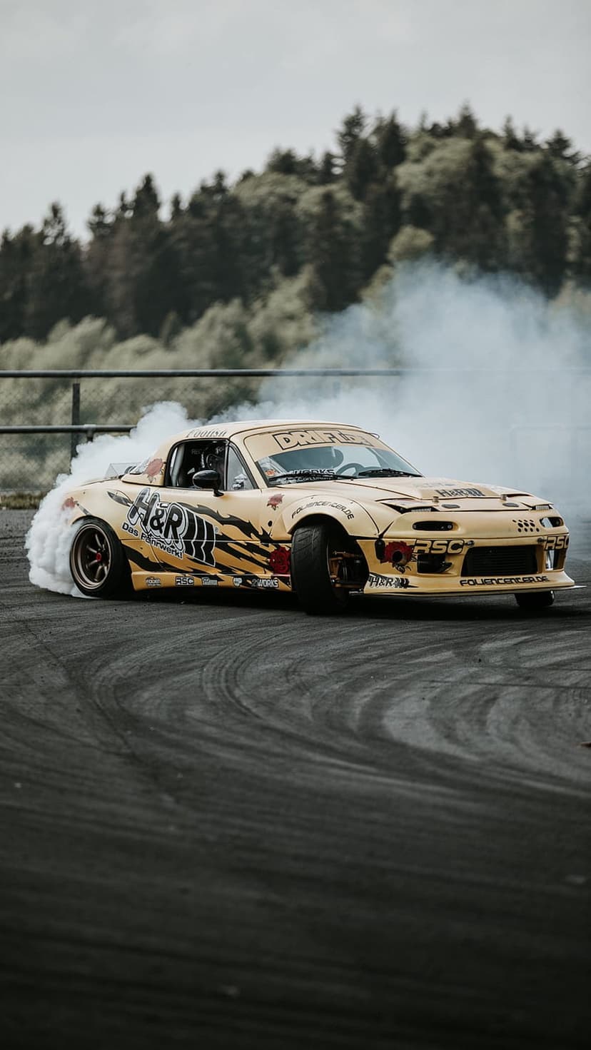 Yellow Mazda MX-5 drift car kicks up white smoke on a race track with a blurred forest and grey sky background.