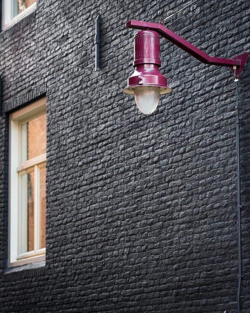 Purple vintage street lamp mounted on a textured black brick wall beside a cream window frame in vertical view.