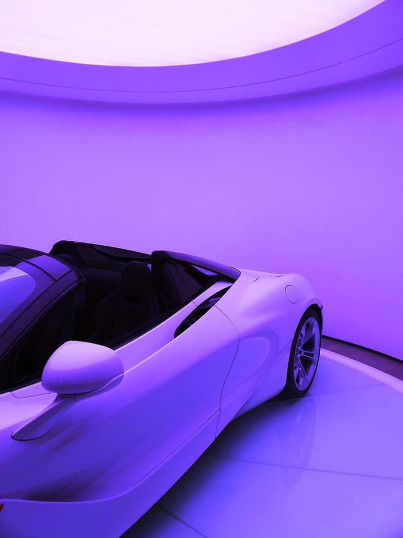 White convertible sports car on a reflective floor illuminated by vibrant purple neon lighting in a dark room.